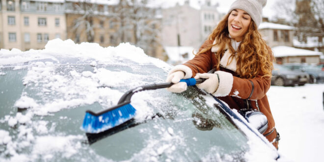Junge Frau reinigt im Winter mit einem Schneebesen die vereiste Windschutzscheibe ihres Autos.